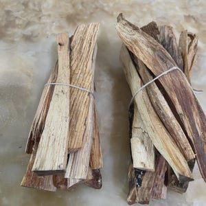 May include: Two bundles of light brown, splintered wooden sticks, each tied together with a thin, white string. The sticks have a rough, fibrous texture with visible grain patterns and varying shades of tan and dark brown throughout the wood pieces.