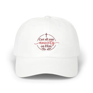 May include: A white baseball cap featuring a circular maroon graphic on the front. The design includes a small cross at the top, the text "Cast all your anxiety on Him." in a mix of serif and script fonts, and decorative leaves at the bottom.