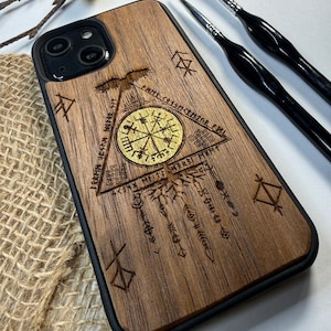 Op de afbeelding: Een rechthoekig smartphonehoesje met een achterpaneel van donker hout voorzien van een lasergegraveerd ontwerp. De afbeelding bevat een centraal goudkleurig Vegvisir-symbool in een driehoek, omringd door runeninscripties en een vogel.