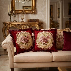 May include: Two square deep red velvet throw pillows with ruffled edges and a central circular tapestry panel featuring a floral bouquet in shades of pink, cream, and green. The text CushionByDimro is printed on the fabric of the right pillow.