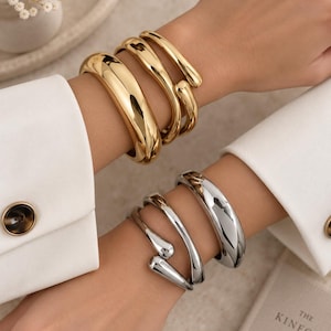 Puede incluir: Un conjunto de tres brazaletes de metal en tono dorado con un acabado liso y pulido en una muñeca, y un conjunto de tres brazaletes de metal en tono plateado con un acabado liso y pulido en la otra, ambos con diseños abiertos.