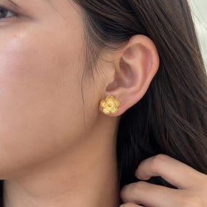 Puede incluir: Un pendiente de botón dorado con forma de flor de cinco pétalos. El pendiente presenta un patrón de líneas radiantes texturizadas en cada pétalo y una pequeña cuenta dorada lisa y elevada en el centro de la flor, sujeto al lóbulo.