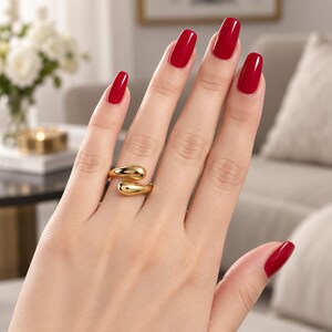 Puede incluir: Un anillo dorado con diseño de bypass que presenta dos extremos lisos, redondeados y en forma de gota. El anillo se lleva en un dedo con esmalte de uñas rojo brillante, destacando el acabado metálico pulido y el estilo abierto y ajustable.