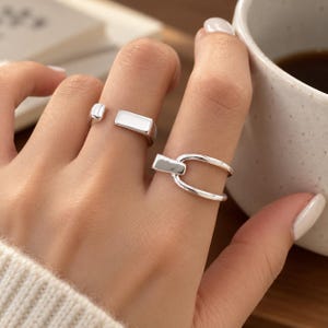 Puede incluir: Dos anillos de tono plateado usados en los dedos. Un anillo presenta una banda abierta simple con un extremo de barra rectangular plana. El segundo anillo tiene un diseño en forma de U con un acabado liso y pulido, creando una apariencia metálica limpia y reflectante.