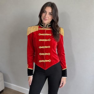 May include: A red velvet jacket featuring gold braided cord closures, gold fringe epaulets on the shoulders, and black velvet trim on the high collar and cuffs. The jacket is worn with plain black trousers.