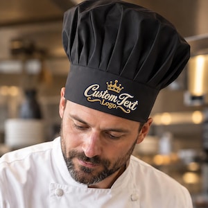 Könnte beinhalten: Eine schwarze Kochmütze mit dem gestickten goldenen Text Custom Text Logo und einem kleinen Kronensymbol darüber. Die Mütze hat ein gefaltetes Oberteil und ein strukturiertes Band. Der Koch trägt eine weiße zweireihige Jacke in einer Profiküche.