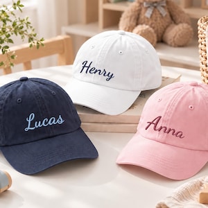 Könnte beinhalten: Drei Baseballkappen in Marineblau, Weiß und Hellrosa. Jede Kappe ist vorne mit gesticktem Text versehen: Die marineblaue Kappe zeigt Lucas in Hellblau, die weiße Kappe Henry in Dunkelblau und die rosa Kappe Anna in Dunkelrosa.