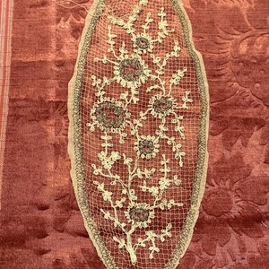 以下が含まれることがあります： クリーム色とダークブラウンの糸で複雑な花柄が刺繍された、楕円形のベージュのレース。中央の茎から枝分かれした蔓と、いくつかの円形の花のモチーフが、暗い色の縁取りで囲まれているデザインです。