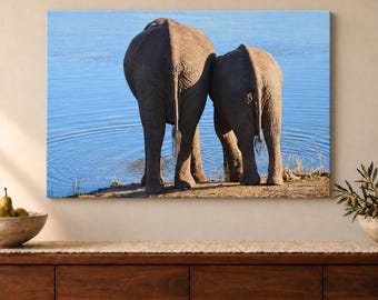 May include: A rectangular wall art print featuring a rear view of two gray elephants standing side by side at the edge of a blue body of water. The elephants are positioned on a sandy bank, with their tails hanging down toward the ground.