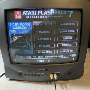 May include: A black Daewoo television displaying the Atari Flashback 7 classic game console menu. The screen shows a list of games including 3-D Tic-Tac-Toe, Adventure, and Asteroids, with yellow and white RCA cables plugged into the front panel.