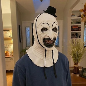 Puede incluir: Una máscara de payaso blanca con un sombrero de copa negro, cejas pintadas de negro y una amplia sonrisa con dientes. La máscara cuenta con un cuello blanco tipo capucha con lazos negros colgantes, sobre una camiseta azul lisa de manga larga.