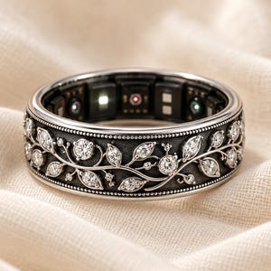 May include: A silver-toned smart ring featuring an intricate, dark-finished band with a raised vine and leaf pattern set with clear, round-cut stones. The interior of the ring displays visible electronic sensors and small LED lights.