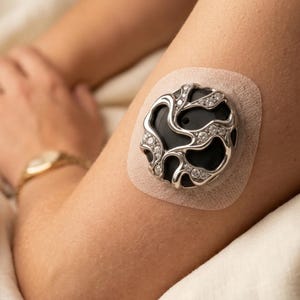 May include: A circular medical sensor cover featuring a black center and a silver-toned, organic-shaped overlay adorned with small, clear, sparkling stones. The device is attached to the skin with a beige, adhesive, fabric-like patch.