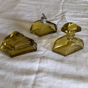 Pode incluir: Três objetos de vidro verde-oliva consistindo em uma caixa com tampa curva, um frasco pequeno com bico pulverizador de metal prateado e um decantador com tampa de vidro geométrica correspondente, todos com superfícies lisas e translúcidas.