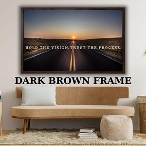 May include: A rectangular wall art piece featuring a dark brown frame surrounding a landscape photograph of a road stretching toward a sunset. The image includes the white text, HOLD THE VISION, TRUST THE PROCESS, centered over the road.