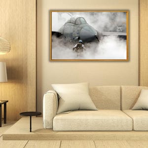 May include: A framed wall art print featuring a gray fighter jet on an aircraft carrier deck surrounded by thick steam. The jet displays the number 101 on its side. A crew member in a vest stands in front of the aircraft amidst the dense vapor.