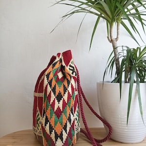Può includere: Uno zaino con chiusura a coulisse caratterizzato da un motivo tessile intrecciato nei toni del rosso scuro, arancione, verde, nero e crema. La borsa presenta spessi cordini intrecciati color bordeaux e una chiusura arricciata superiore.