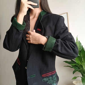 May include: A black jacket featuring a dark green stand-up collar and matching green cuffs. The garment includes red-trimmed pockets and intricate green embroidery detailing near the lower front sections, with a button-down front closure.