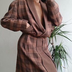 May include: A long-sleeved, mid-length wrap dress in a brown and dark brown plaid pattern. The garment features a shawl collar, a deep V-neckline, and a tie-waist closure. The fabric has a textured, knit-like appearance with structured shoulders.