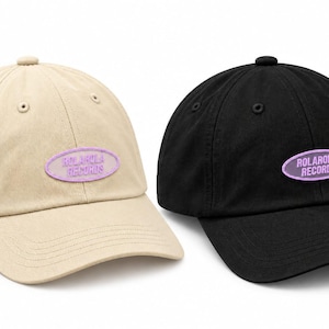 May include: Two baseball caps, one beige and one black, each featuring a centered oval patch with the text ROLAROLA RECORDS in purple. Both caps have a structured crown, a curved brim, and matching top buttons, showcasing a classic six-panel design.