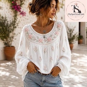 May include: A white long-sleeved blouse featuring a round neckline with intricate pink floral embroidery and green leaf accents. The garment has a loose fit, gathered cuffs, and scattered floral embroidery patterns across the front and sleeves.