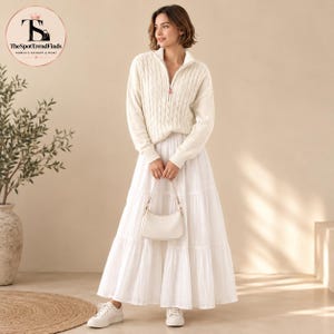 May include: A cream-colored cable knit sweater with a quarter-zip collar paired with a long, white, tiered maxi skirt. The outfit is accessorized with a small, solid white shoulder bag and white sneakers, creating a monochromatic look.