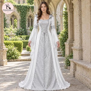 May include: A floor-length gown featuring a silver patterned center panel and white side panels with long flared sleeves. The bodice includes a lace-up corset detail with a central tie, a square neckline, and a hood. The fabric has a subtle sheen.