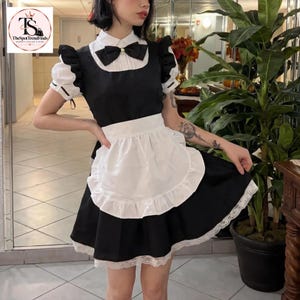 May include: A black and white dress featuring a white collared bodice with a black bow tie, short puffed sleeves with black trim, and a white apron with a ruffled edge. The skirt is black with a white lace trim along the bottom hemline.