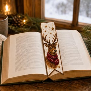 May include: A rectangular bookmark featuring a beaded illustration of a brown deer with large antlers wearing a red textured scarf. The background of the bookmark is cream-colored with small gold star accents and a pointed bottom edge.