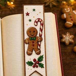 May include: A rectangular cross-stitch bookmark on white fabric with a brown border. The design features a brown gingerbread person holding a red and white candy cane, green holly leaves with red berries, and small red and green star accents.