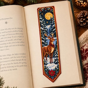 May include: A rectangular cross-stitch bookmark featuring a brown deer standing in a snowy forest at night. The design includes a yellow full moon, dark blue sky, evergreen trees, and a red poinsettia flower at the bottom, all bordered in red.