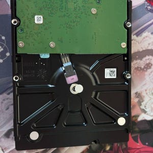 Peut inclure: Un disque dur noir doté d'une carte de circuit imprimé verte bien visible sur sa partie supérieure. L'appareil comprend des vis de montage argentées, un connecteur d'interface SATA en haut et des étiquettes blanches avec des codes-barres.