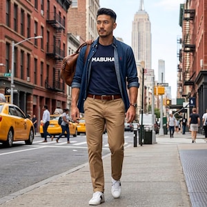 May include: A navy blue t-shirt with the white text MABUHA, worn under an unbuttoned blue denim shirt. The outfit includes tan chinos, a brown leather belt, white sneakers, a brown leather bag, and a wristwatch, set against a city street background.