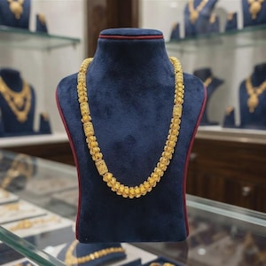 Puede incluir: Un collar de cadena de oro amarillo con un patrón repetitivo de eslabones cilíndricos texturizados y cuentas esféricas más pequeñas. El collar se exhibe sobre un busto de terciopelo azul oscuro, mostrando un acabado metálico pulido.