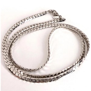 May include: A silver-toned chain necklace featuring a flat, interlocking herringbone link pattern. The necklace includes a lobster claw clasp and a small rectangular metal tag engraved with the brand name MONET near the closure mechanism.
