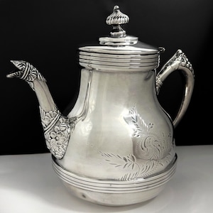 May include: A polished silver teapot featuring a rounded body with engraved floral and leaf patterns. The spout is shaped like an animal head with detailed carvings at the base. It includes a matching lid with a finial and a curved handle.