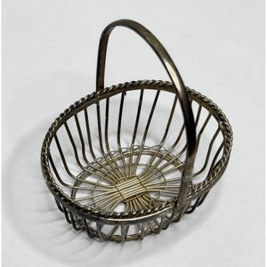 May include: A silver-toned metal basket featuring a rounded, open-wire design with a twisted rope-style rim and a single arched handle. The base is constructed from a series of thin, radiating metal wires that converge in a rectangular center.