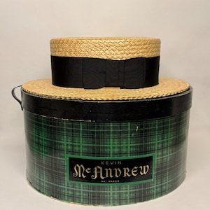 May include: A straw boater hat with a black ribbon band resting on a round, green and black plaid hat box. The box features a black label with the text Kevin McAndrew Hat Maker in gold lettering, centered on the front of the cylindrical container.