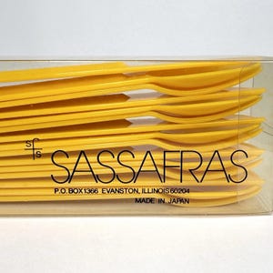 May include: A set of bright yellow plastic spoons stacked inside a clear rectangular box. The packaging features the text SASSAFRAS, the logo sfs, the address P.O. BOX 1366 EVANSTON, ILLINOIS 60204, and the text MADE IN JAPAN printed on the front.
