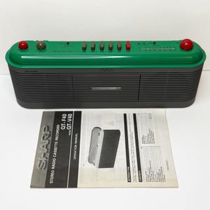 Peut inclure: Un magnétophone radio stéréo Sharp avec un panneau supérieur vert doté de boutons et de molettes rouges et gris, et un boîtier de haut-parleur nervuré gris foncé. Un manuel d'utilisation pour les modèles QT-F40 et QT-V40 est placé devant.