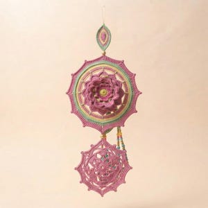 May include: A decorative hanging ornament featuring two crocheted mandalas in shades of pink, yellow, and light green. The top mandala includes a central three-dimensional flower, while the bottom piece is flat. Small beads accent the structure.
