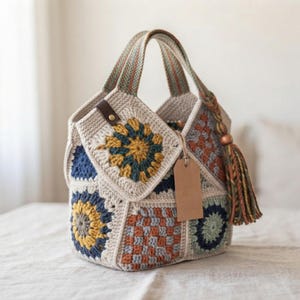 Può includere: Una borsa all'uncinetto composta da vari motivi quadrati e circolari nei colori crema, giallo senape, blu navy e arancione bruciato. La borsa presenta due manici integrati, una linguetta in pelle marrone con rivetto e una grande nappa in filato.