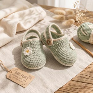 May include: A pair of sage green crocheted baby clogs featuring a small white daisy appliqué with a yellow center on each shoe. Each clog has a brown wooden button on the side strap. A tan paper tag reads BABY CLOGS CROCHET PATTERN PDF DOWNLOAD.