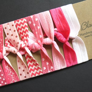 Könnte beinhalten: Ein Set aus pinken und weißen Haargummis mit verschiedenen Mustern, darunter Punkte, Herzen und Zickzackmuster. Die Haargummis sind fächerförmig auf einem braunen Hintergrund angeordnet. Der Text "Bloomzberry Handmade Hair Accessories www.bloomzberry.etsy.com" ist auf den Hintergrund gedruckt.
