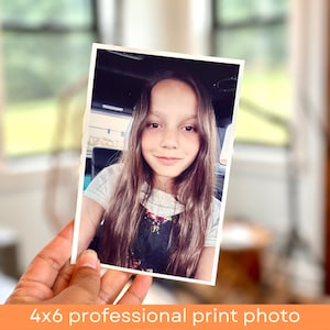 May include: A 4x6 inch professional print photo of a young girl with long brown hair, wearing a white shirt and a floral print overall dress. She is smiling and looking at the camera.