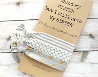 May include: A card with the text "I found my MISTER But I still need My SISTER Will you be my bridesmaid?" with a set of three hair ties in silver and gold with different patterns.
