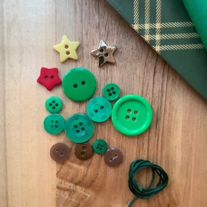 May include: A collection of sewing supplies including green, brown, and yellow buttons in various sizes and shapes, including stars and round buttons. A spool of green thread and green plaid fabric are also present.
