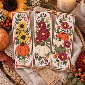 May include: Three rectangular cross-stitch bookmarks featuring autumn motifs. The left shows sunflowers and an orange pumpkin, the centre displays deep red flowers and a white pumpkin, and the right features orange and white pumpkins with flowers.