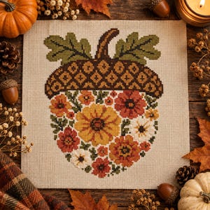 May include: A square piece of beige fabric featuring a cross-stitch design of an acorn. The acorn cap is brown with a dark grid pattern and green leaves. The base of the acorn is filled with a colorful arrangement of orange, yellow, and white flowers.