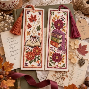 May include: Two rectangular cross-stitch bookmarks on a cream fabric base. The left bookmark features a steaming mug on an open book with orange flowers and maple leaves. The right bookmark shows a stack of three books with a purple scarf and flowers.